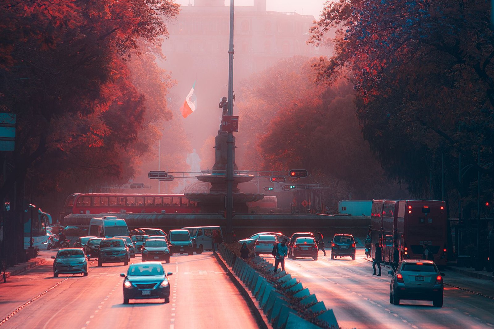 Avenida con tráfico y un monumento con bandera mexicana al fondo en luz cálida de atardecer, personas a la distancia sin rostro distinguible