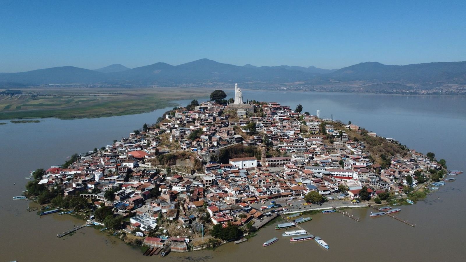Vista aérea de la isla de Janitzio con sus casas y una estatua en la cima, rodeada por el lago de Pátzcuaro y cerros al fondo, sin personas en primer plano