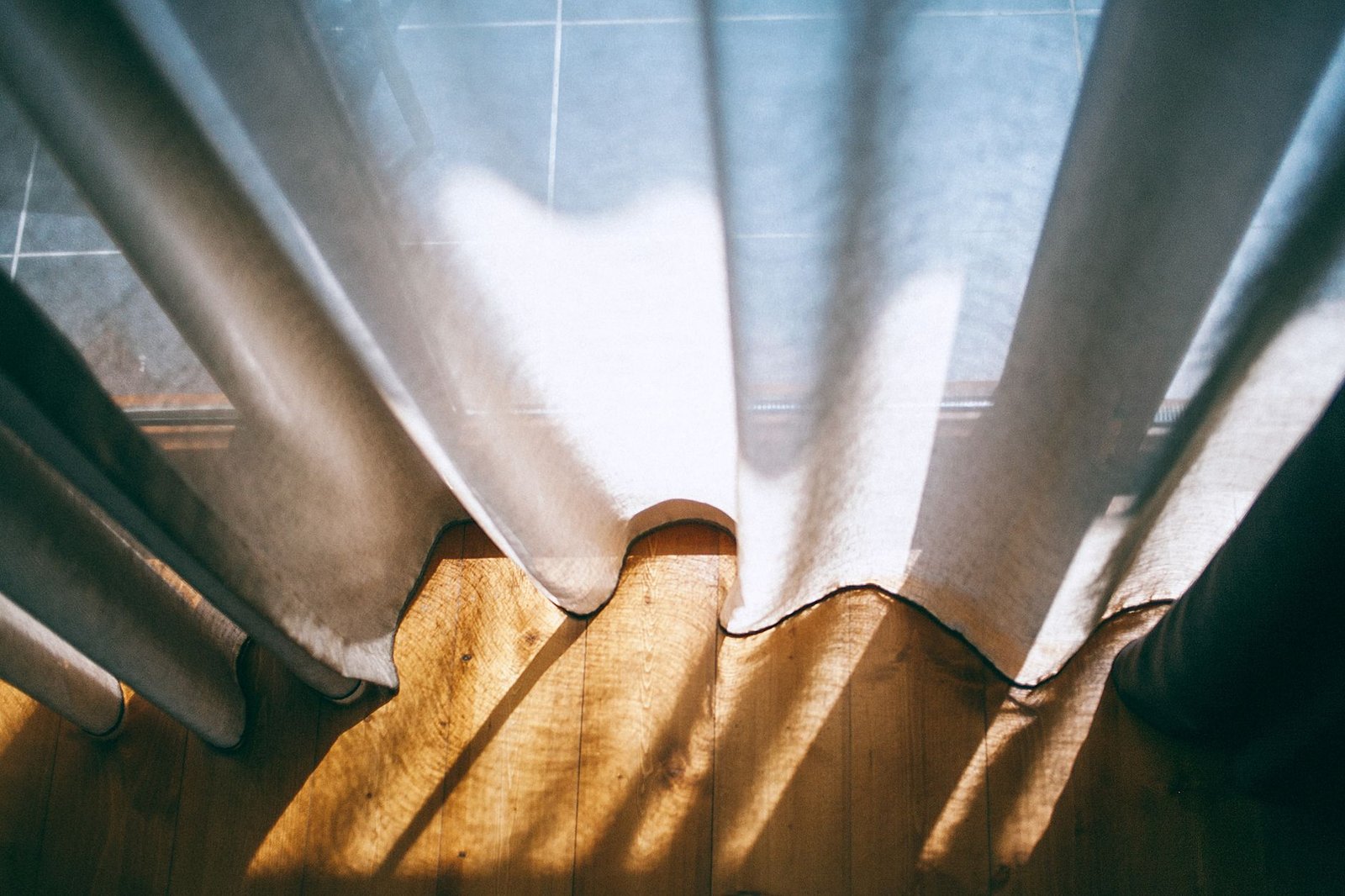 Una cortina blanca semitransparente filtra luz de sol cálida sobre un piso de madera, en un espacio privado y en calma
