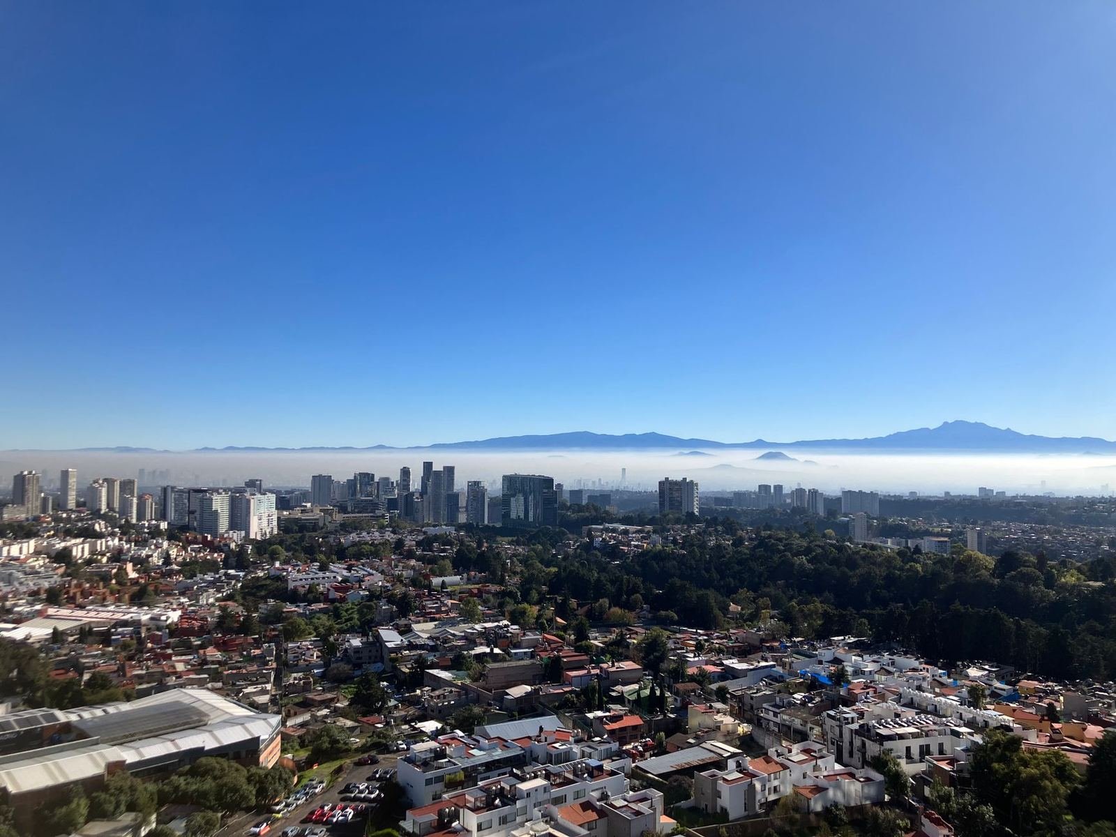 Vista aérea de una zona urbana con árboles y edificios bajo un cielo azul, con montañas al fondo entre la neblina, sin personas ni texto